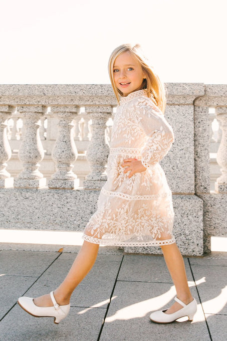 A young girl with blonde hair confidently walks outdoors in sunlight beside a stone balustrade, wearing the Ivy City Co Mini Sicily Dress with Long Sleeves and white shoes.