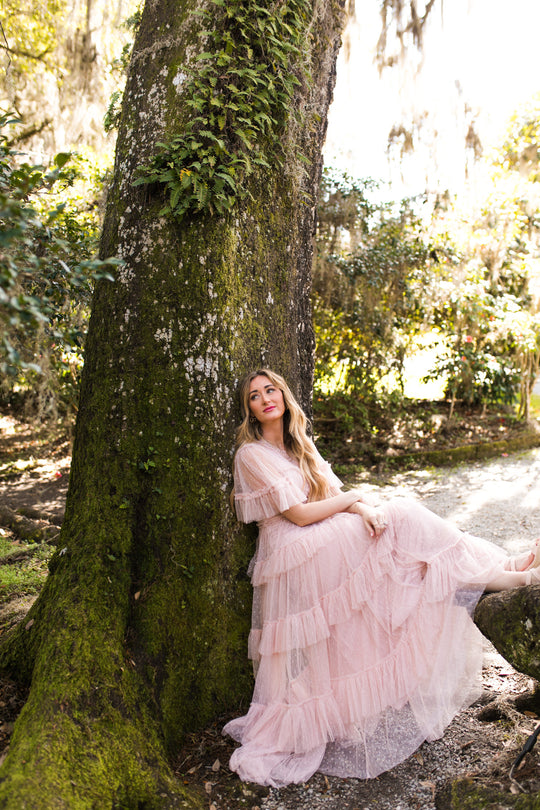 A woman wearing the Whimsical Dress in Blush by Ivy City Co sits thoughtfully against a mossy tree trunk, bathed in dappled sunlight and surrounded by lush greenery.