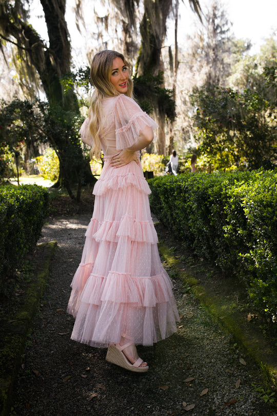 A woman wears the Whimsical Dress in Blush by Ivy City Co, paired with wedge sandals, as she stands on a garden path surrounded by lush greenery and trees in a vibrant outdoor setting.