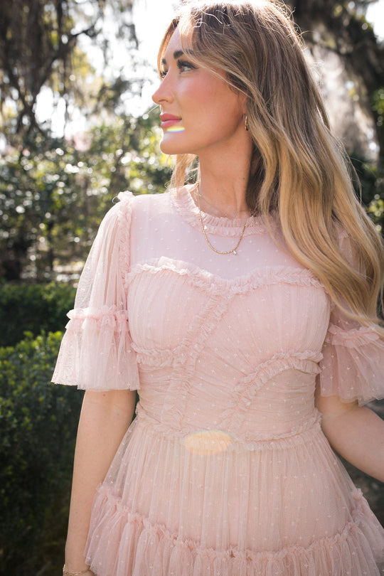 A woman with long blonde hair stands outdoors in sunlight, wearing the Ivy City Co Whimsical Dress in Blush with short sleeves and ruffles. Trees and greenery are blurred in the background.