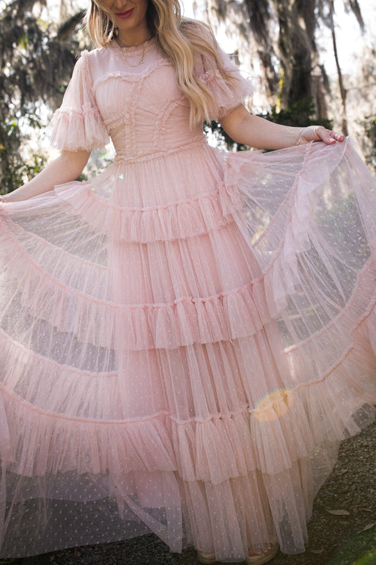 Woman outdoors on a sunny day wearing the Ivy City Co Whimsical Dress in Blush, featuring ruffled, semi-sheer tulle tiers with delicate polka dots, holding out the skirt. Her face is partially out of frame.