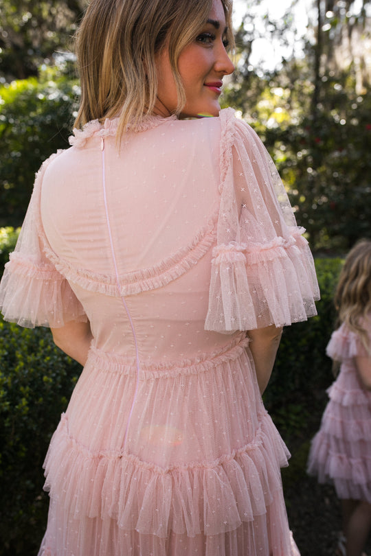 A woman in the Whimsical Dress in Blush by Ivy City Co stands outdoors with her back to the camera, while another girl in a matching dress is seen among greenery and sunlight.