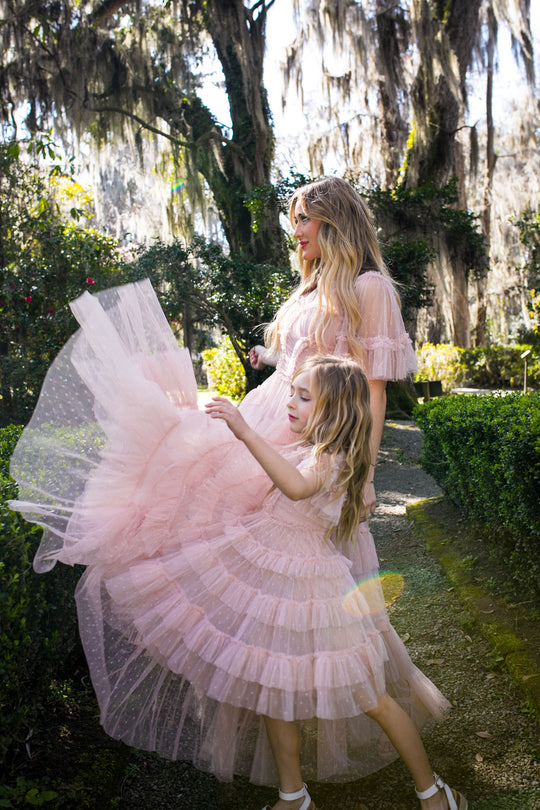 A woman and a young girl wear matching Ivy City Co Whimsical Dress in Blush, strolling along a garden path surrounded by greenery as the girl playfully lifts her dress under the filtered sunlight.