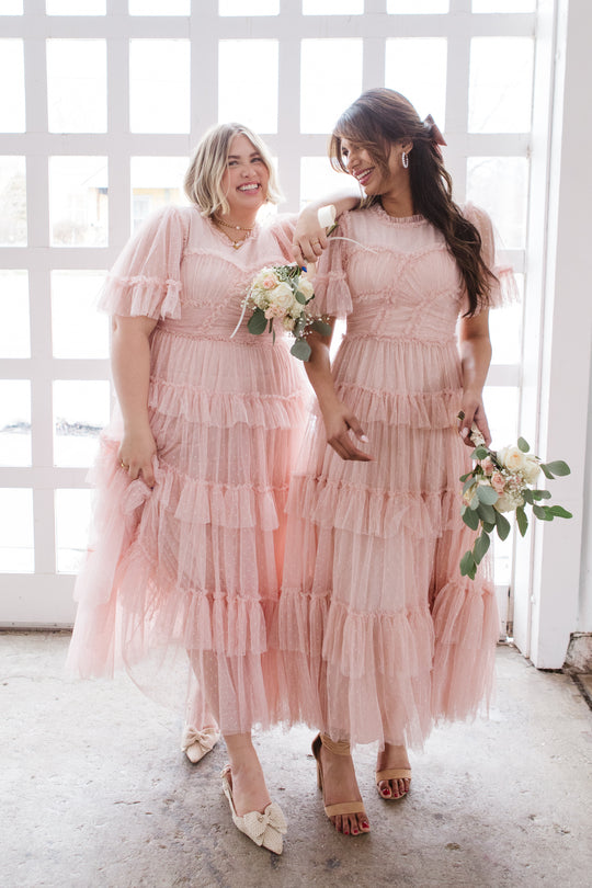 Two women in Ivy City Co's Whimsical Dress in Blush—ruffled tulle skirts—smile before a large window. One has short blonde hair, the other long brown hair with a pink ribbon. Both hold small bouquets.